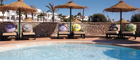 A pool area with lounge chairs and thatched umbrellas, surrounded by a stone wall and palm trees.