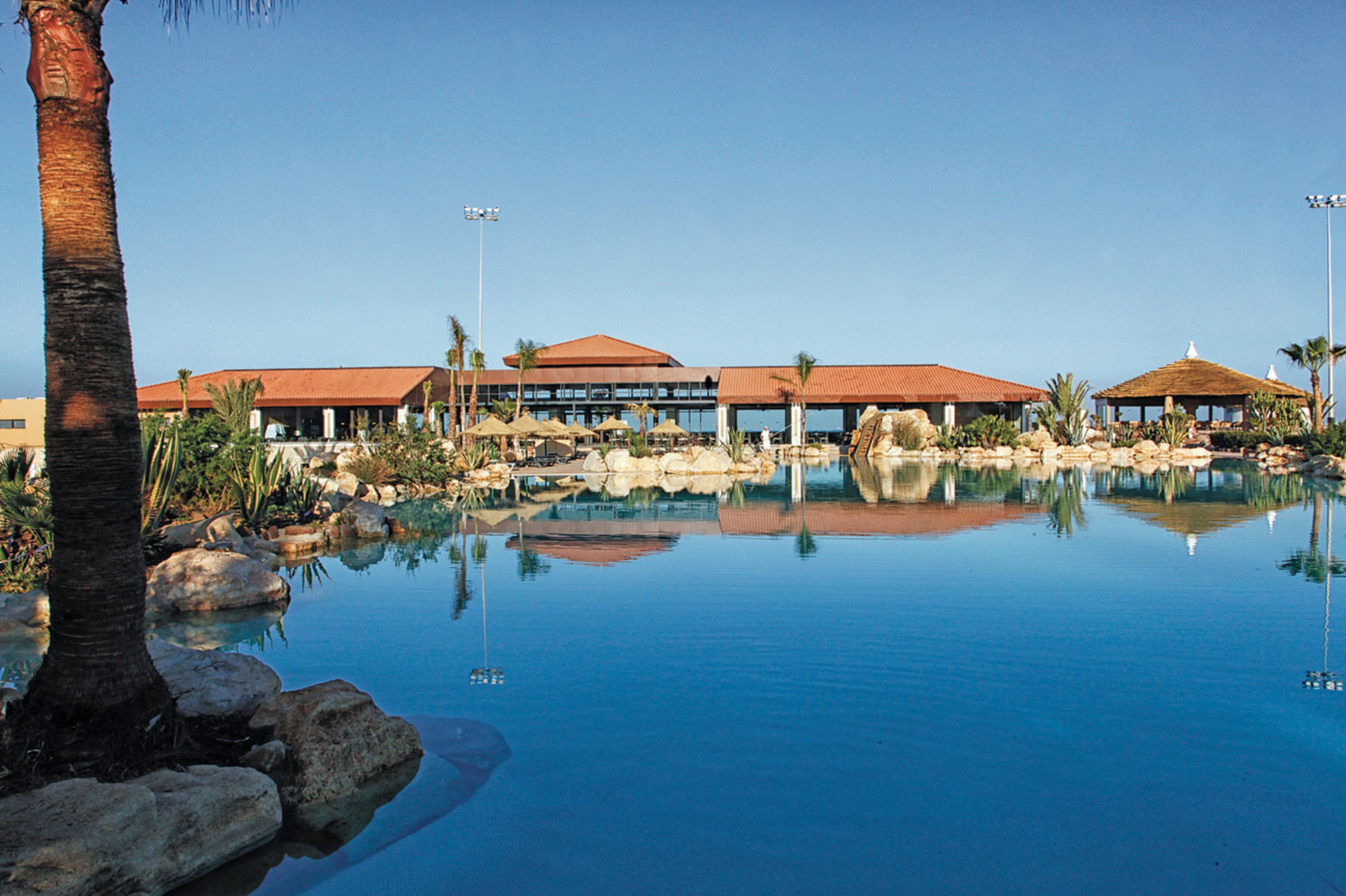 A resort with a clear pool, palm trees, and buildings with red roofs.