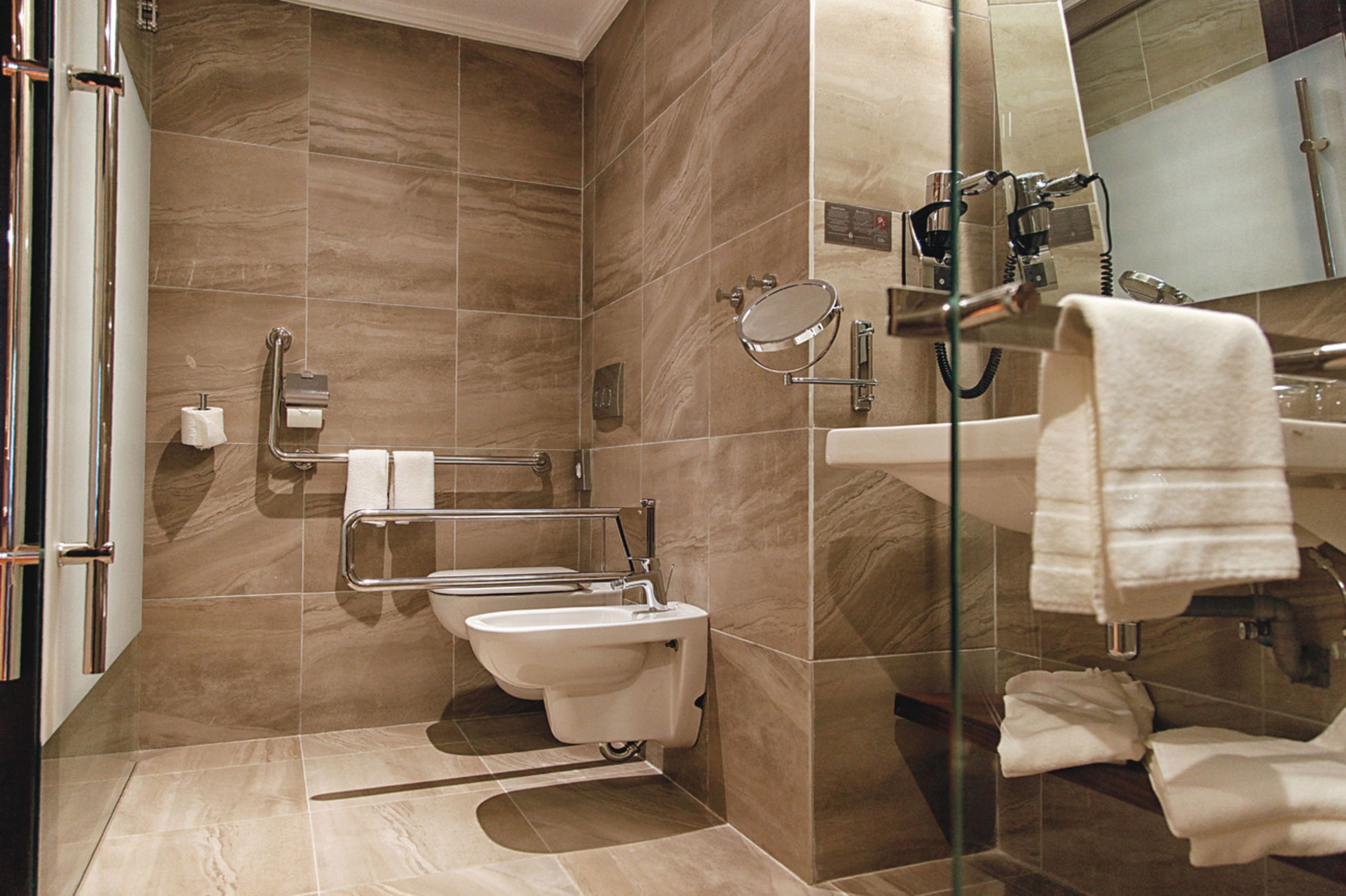 A modern bathroom with a glass shower enclosure, a toilet with a grab bar, a sink with a mirror, and multiple towels.