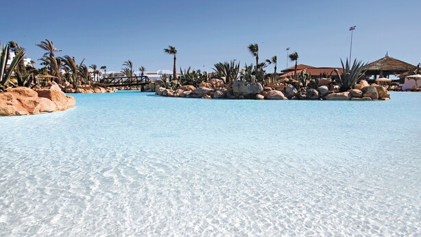 A clear pool with rocks and palm trees, surrounded by buildings. - Hotel Riu Tikida Dunas - All Inclusive (Agadir)