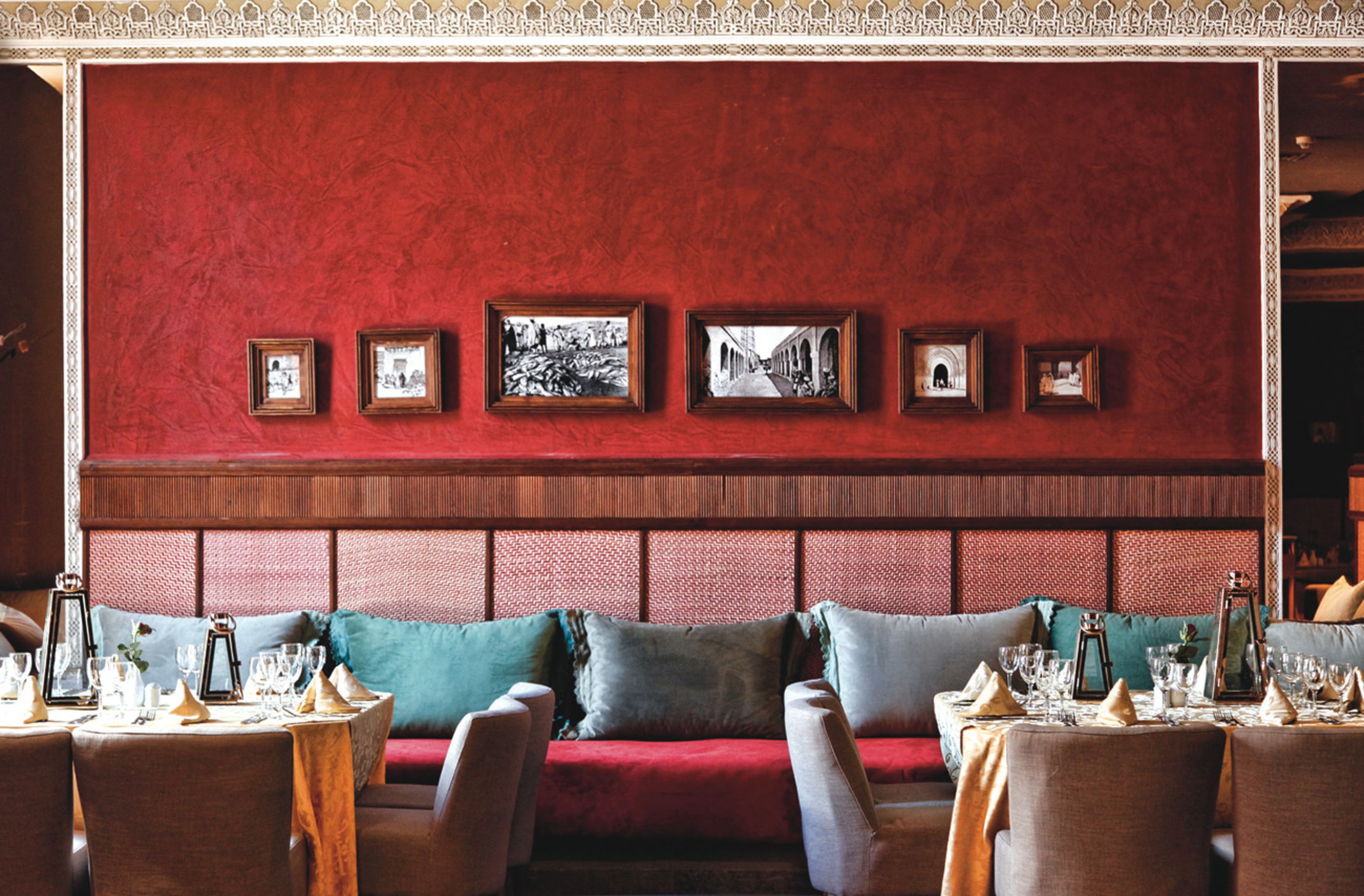 Een restaurantinterieur met een rode bekleden zithoek met een bank, een muur versierd met ingelijste foto's en gedekte tafels.