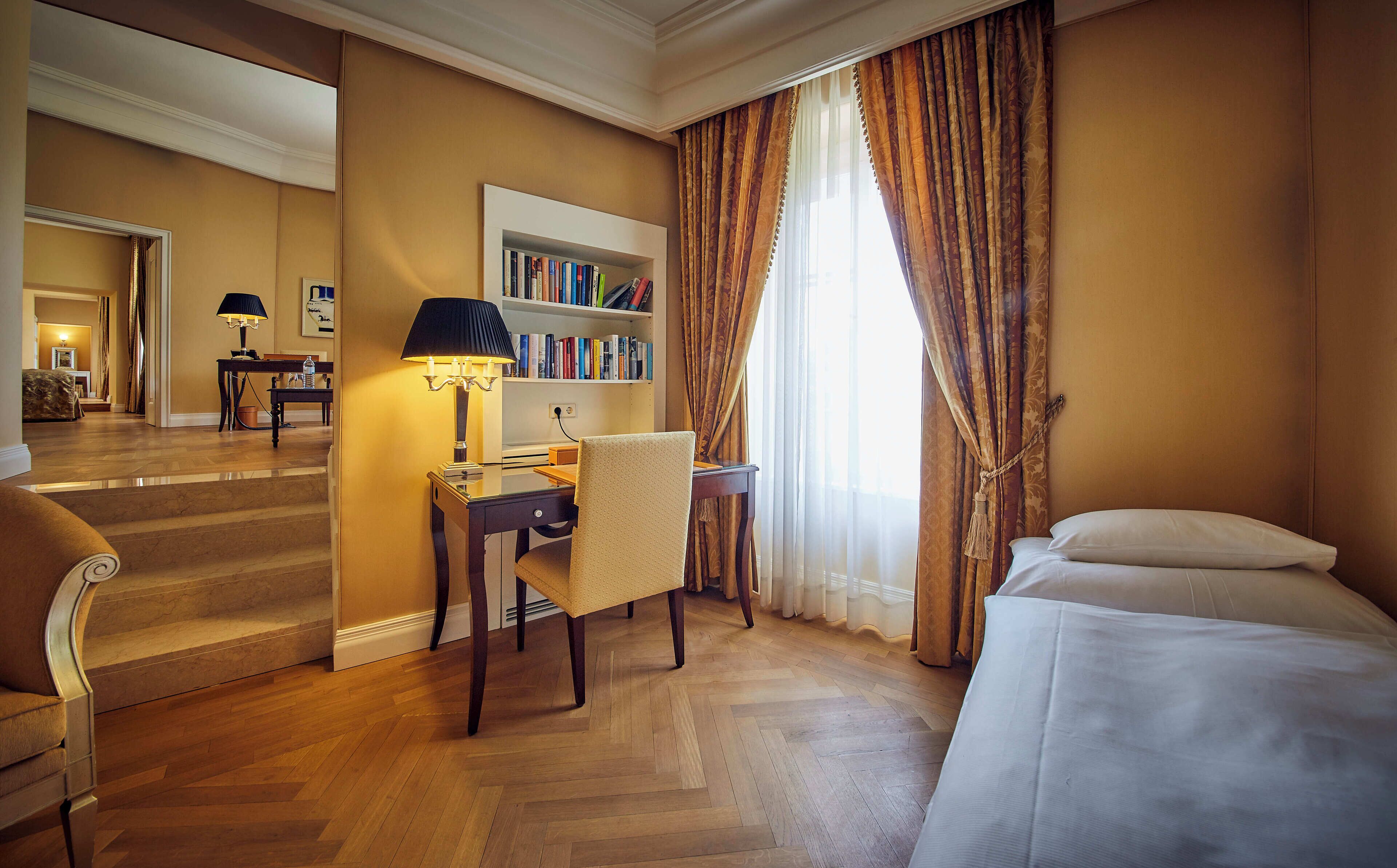 A hotel room with a bed, a desk with a lamp, a chair, a bookshelf, and a staircase leading to another room.