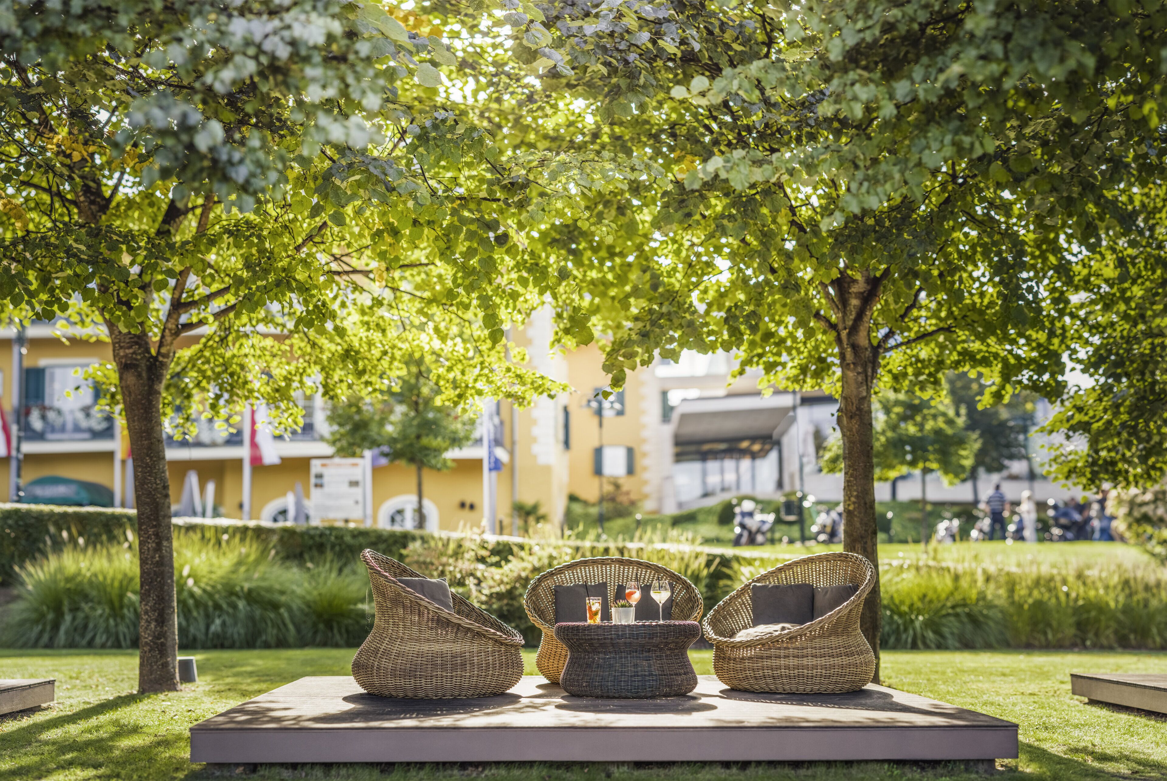 An outdoor seating area with wicker chairs and a table, surrounded by greenery and trees.