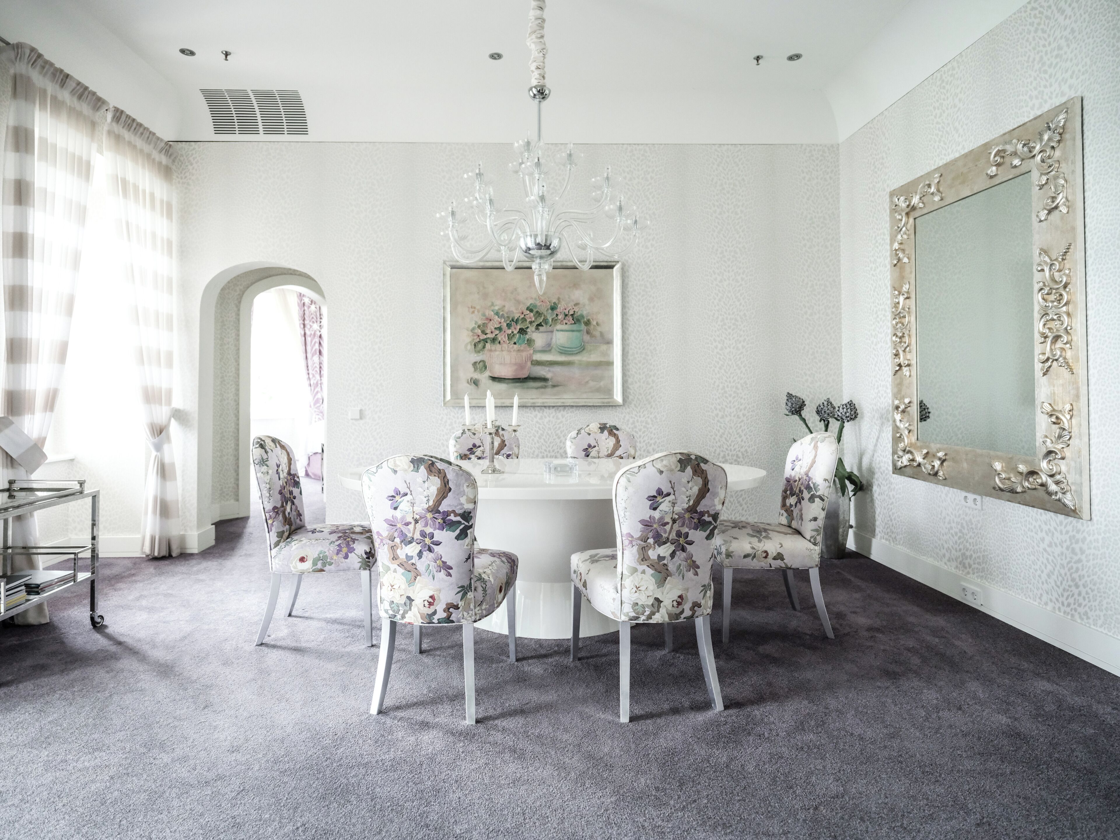 Een eetkamer met een ronde tafel, bloemenstoelen, een kroonluchter, een grote spiegel en een schilderij aan de muur.