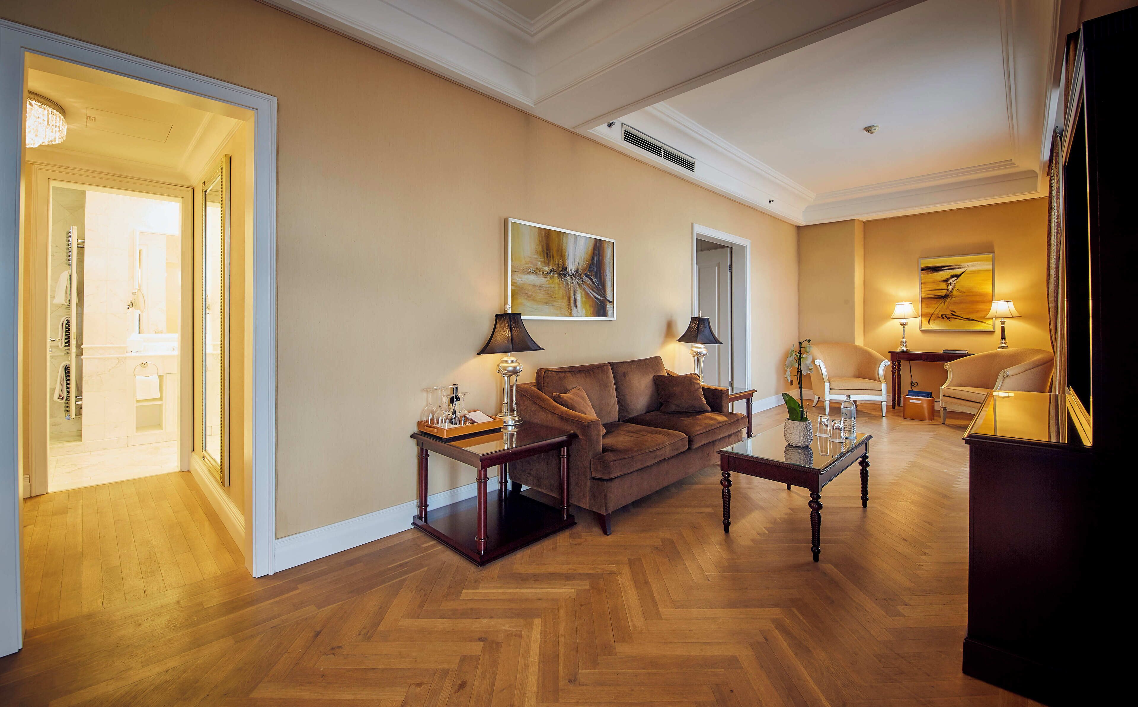 A spacious living room with a brown sofa, two side tables, and a glass coffee table. A door leads to a bathroom, and there are two lamps on the side tables.