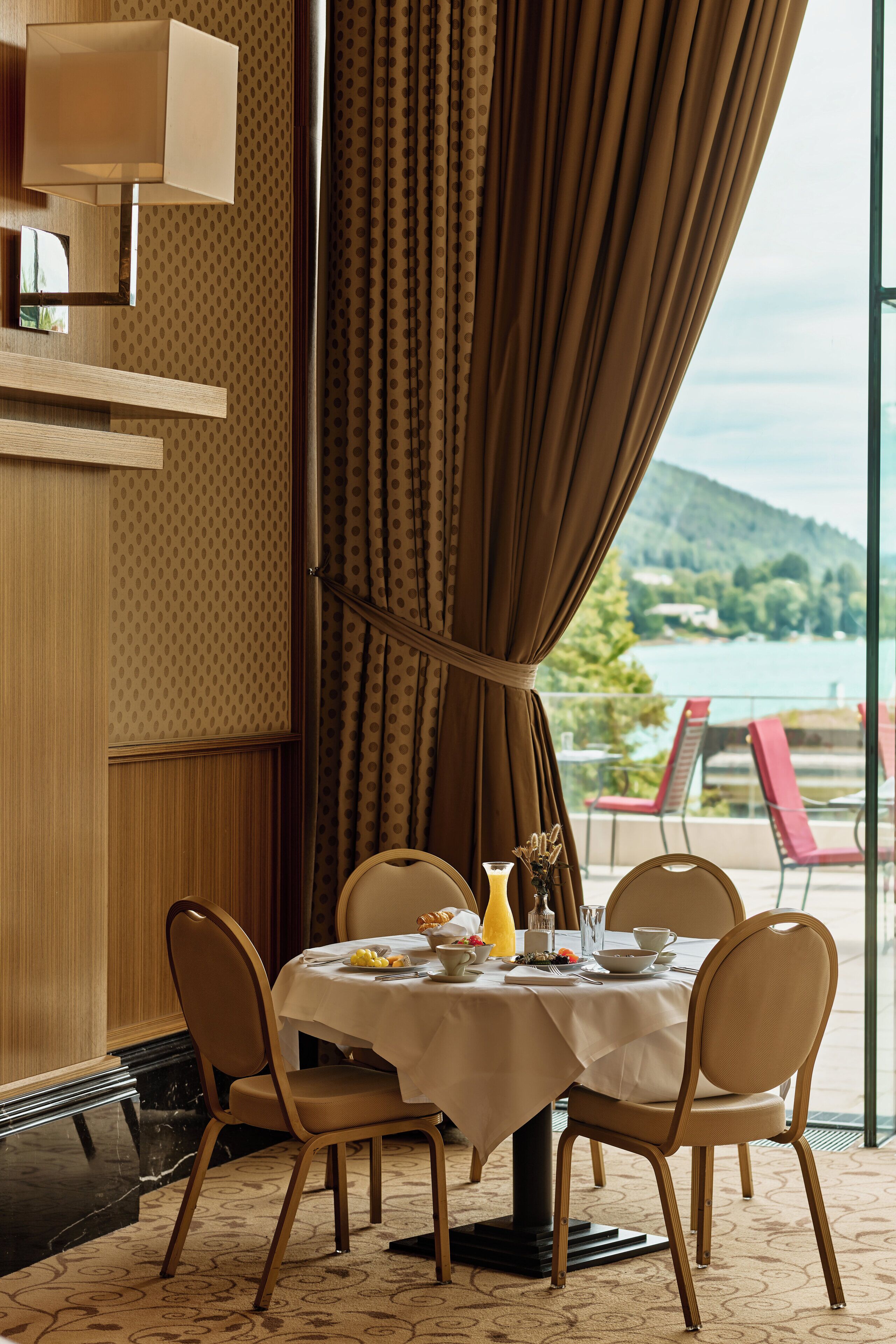 A dining area with a table set for four, chairs, a vase with flowers, and a view of a body of water through large curtains.