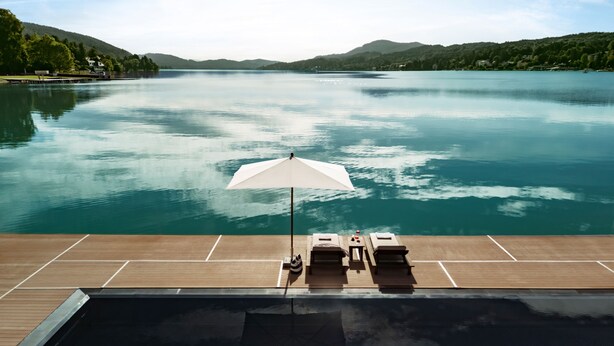 Zona junto a la piscina con vista a un lago y montañas, una sombrilla blanca y sillas de descanso.