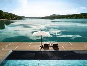 A poolside area with a view of a lake and mountains, a white umbrella, and lounge chairs. - Falkensteiner Schlosshotel Velden – The Leading Hotels of the World (Velden am Wörther See)