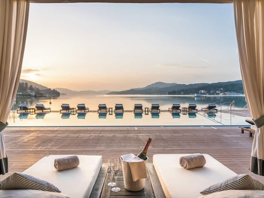 Vista de la piscina con sillas de descanso, una botella de champán y una copa de vino en una terraza de madera con vistas a un cuerpo de agua tranquilo y colinas lejanas.