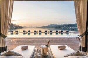 Vista de la piscina con sillas de descanso, una botella de champán y una copa de vino en una terraza de madera con vistas a un cuerpo de agua tranquilo y colinas lejanas.