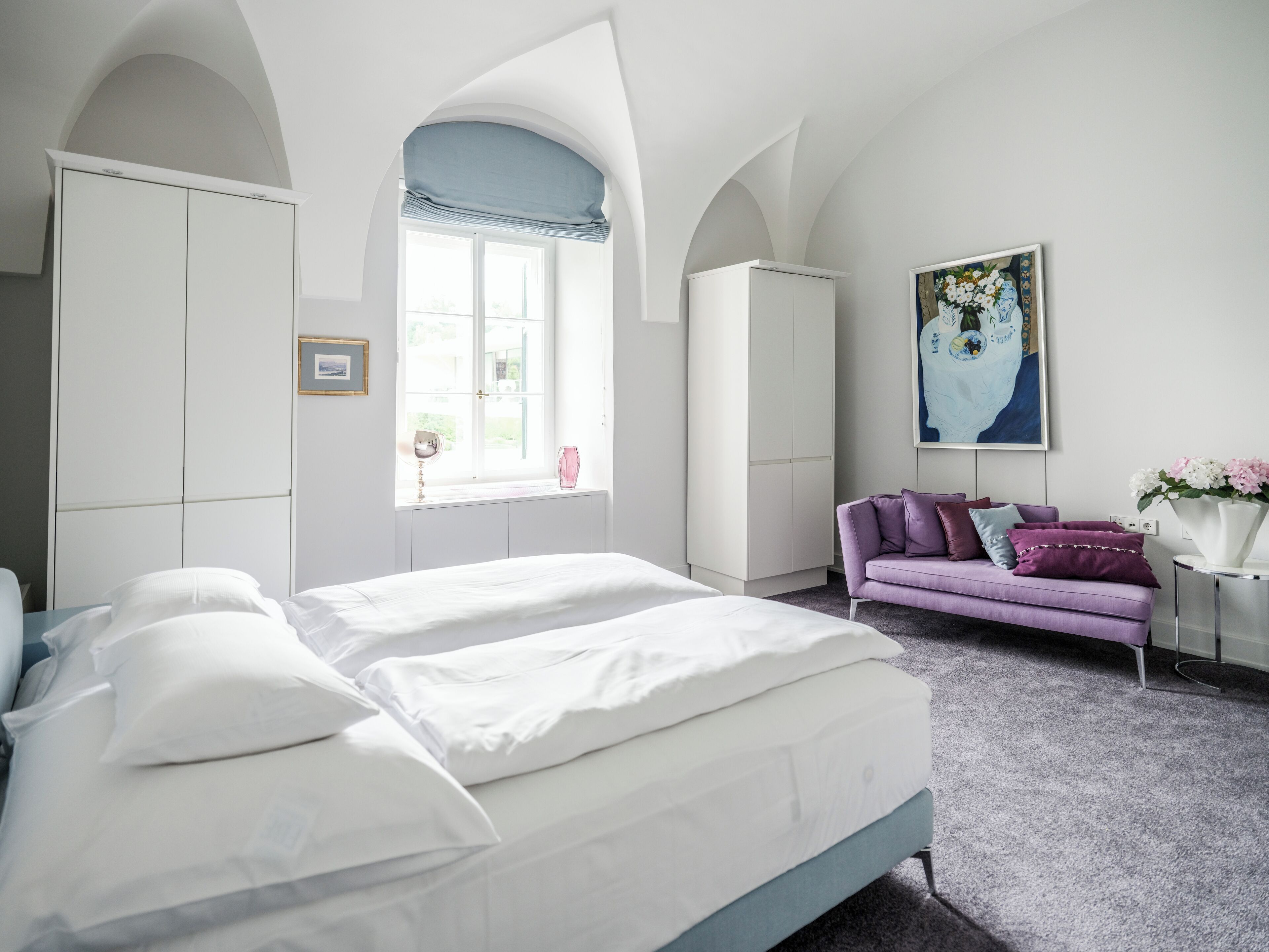A bedroom with a large bed, a purple sofa, and a window with a view of greenery.