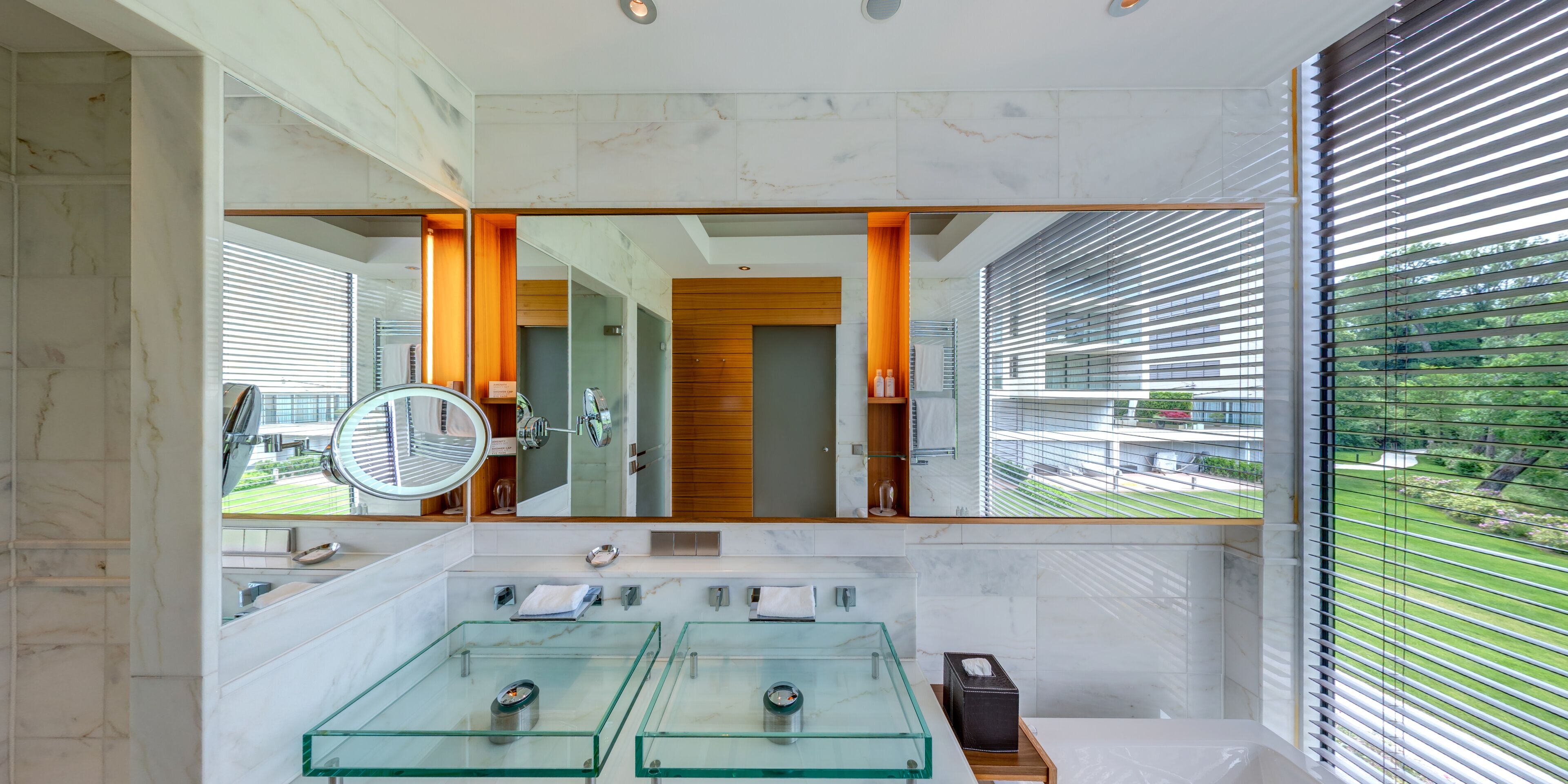 Ein modernes Badezimmer mit einem großen Spiegel, zwei Glaswaschbecken und Blick auf eine grüne Landschaft durch das Fenster.