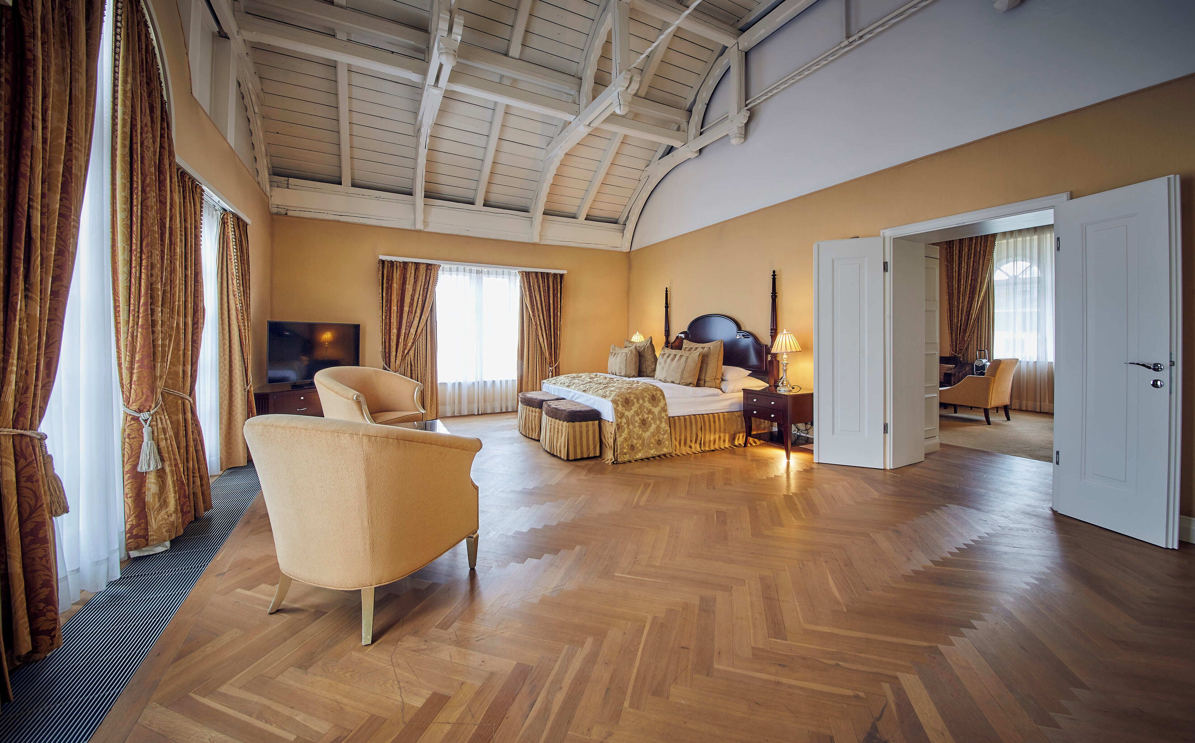 Habitación de hotel espaciosa con una cama grande, un área de descanso con una silla y reposapiés, un televisor y una puerta que conduce a otra habitación.