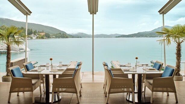 A terrace with a table and chairs overlooking a lake, with mountains in the background. - Falkensteiner Schlosshotel Velden – The Leading Hotels of the World (Velden am Wörther See)
