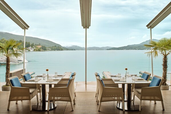 A terrace with a table and chairs overlooking a lake, with mountains in the background. - Falkensteiner Schlosshotel Velden – The Leading Hotels of the World (Velden am Wörther See)
