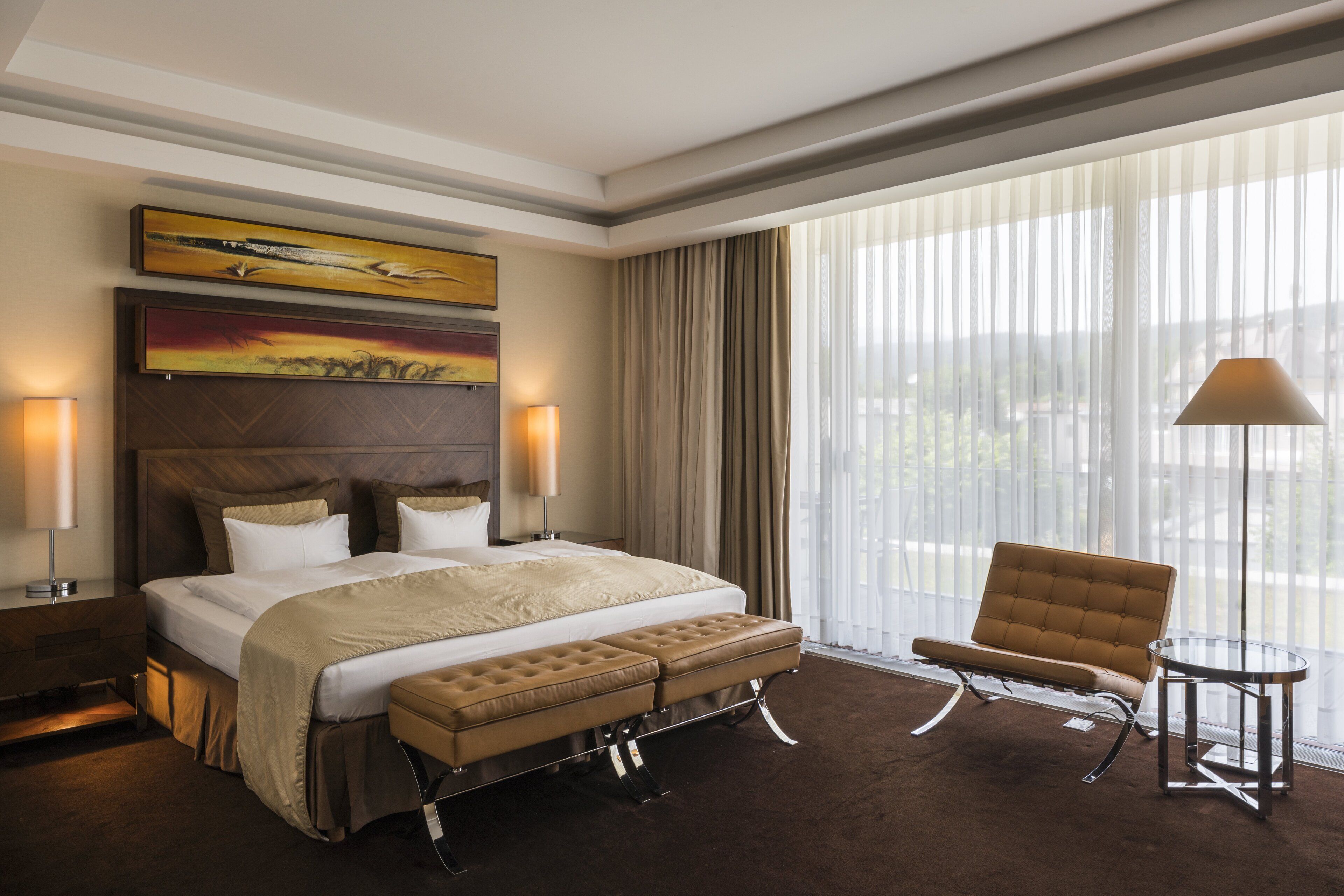 A hotel room with a large bed, a bench, a chair, and a view of the outdoors through sheer curtains.