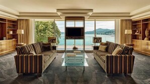 Amplia sala de estar con dos sofás a rayas, una mesa de centro de vidrio y una gran pantalla de televisión. La habitación cuenta con estanterías de madera y vistas a un lago y montañas.