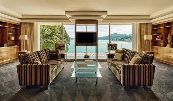 A spacious living room with two striped sofas, a glass coffee table, and a large TV screen. The room features wooden bookshelves and a view of a lake and mountains. - Falkensteiner Schlosshotel Velden – The Leading Hotels of the World (Velden am Wörther See)