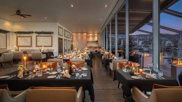 A restaurant with tables set for dining, large windows offering a view of the city, and a ceiling fan.
