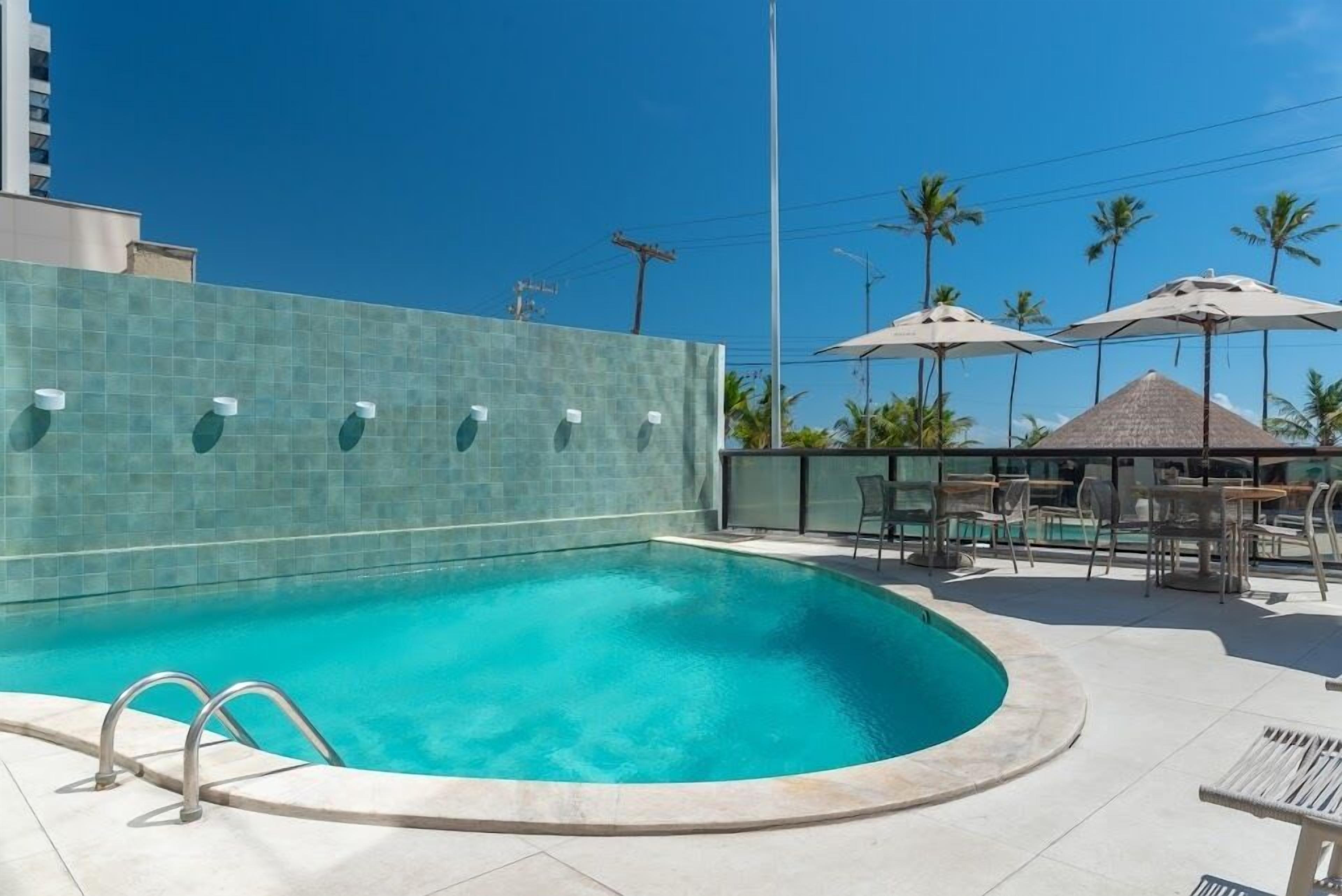 A swimming pool with a curved edge, surrounded by lounge chairs and umbrellas, and a tiled wall in the background.