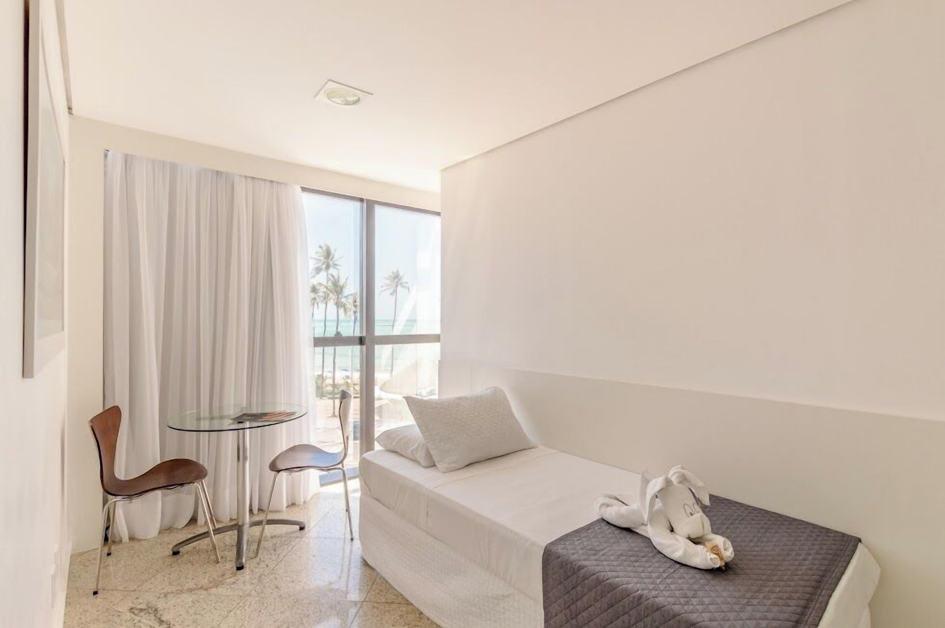 A hotel room with a bed, a small table with chairs, a window with curtains, and a view of palm trees.