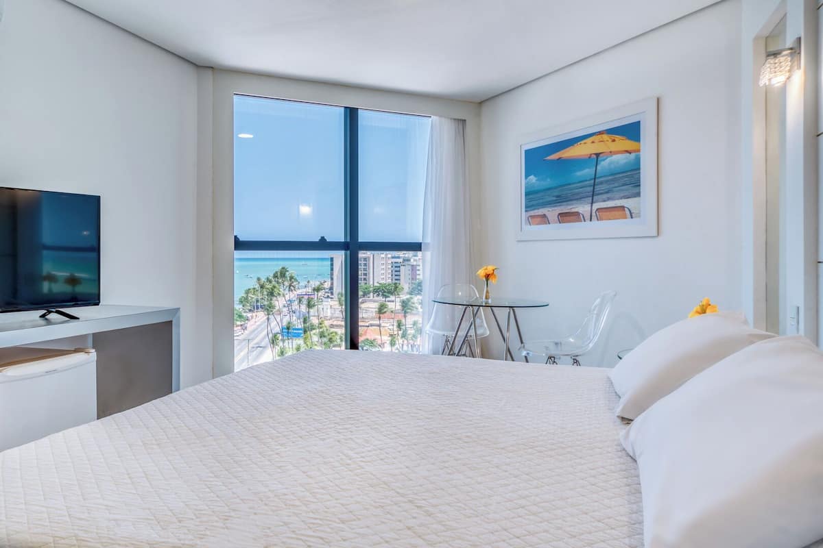 Habitación de hotel con una cama grande, un escritorio con televisor, una mesita con sillas y un amplio ventanal con vistas a la playa y la ciudad.