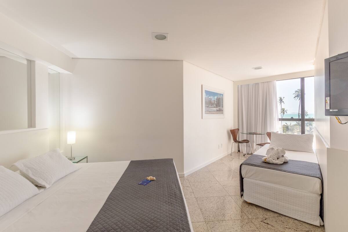 Habitación de hotel con cama, escritorio, silla, televisión y ventana con vista a palmeras.