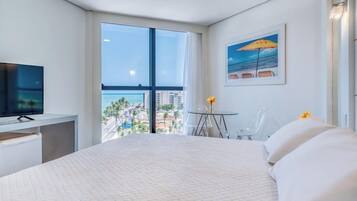 Habitación de hotel con una cama grande, televisión, escritorio, una mesita con un jarrón y un cuadro de una playa.