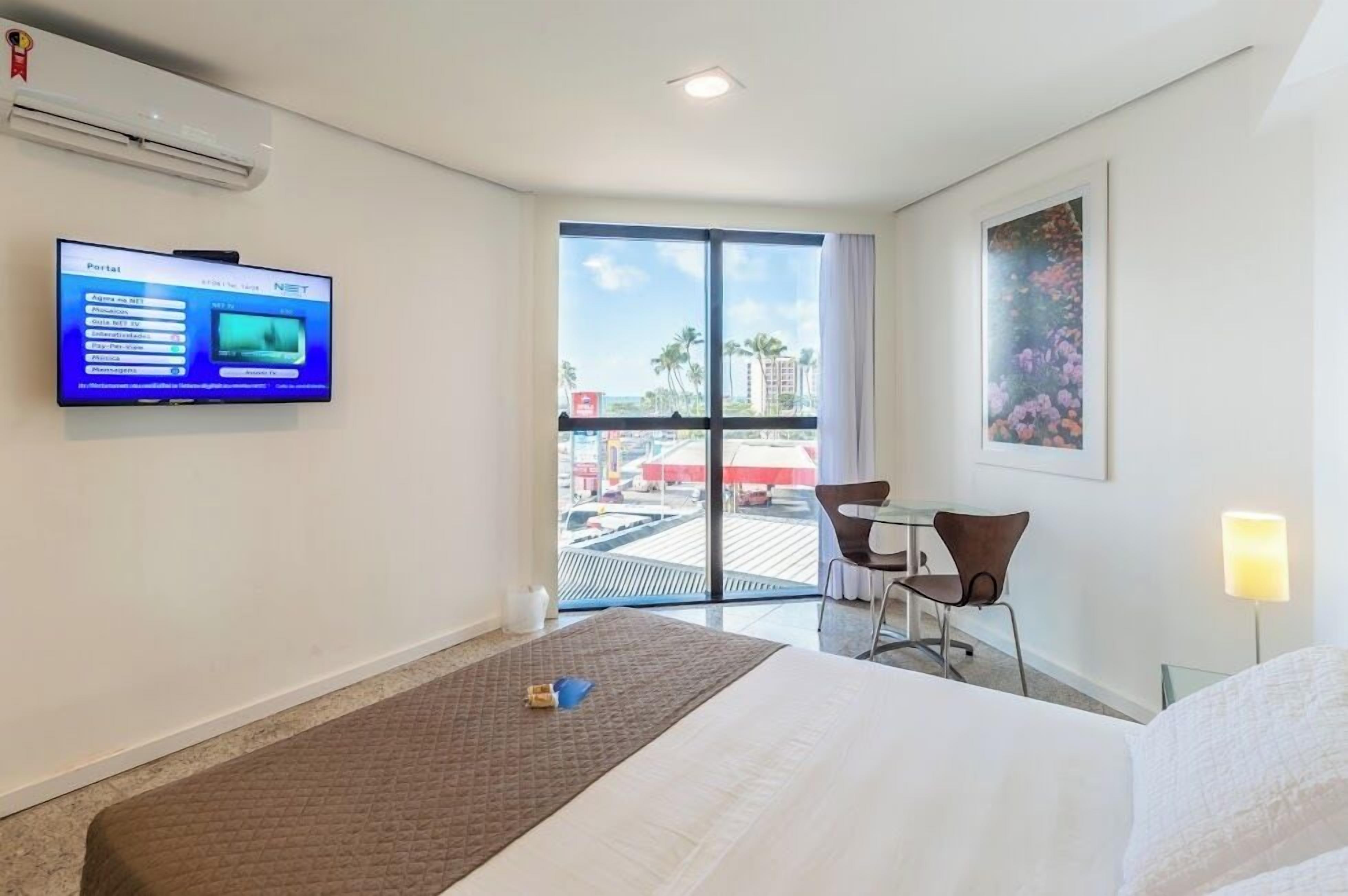 A hotel room with a bed, a desk, a chair, a television, and a large window with a view of palm trees and buildings.