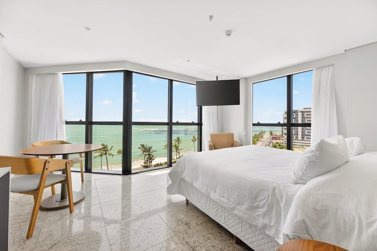 Habitación de hotel con cama, escritorio y silla, ventanas amplias con vista al mar y un televisor de pantalla plana en la pared.