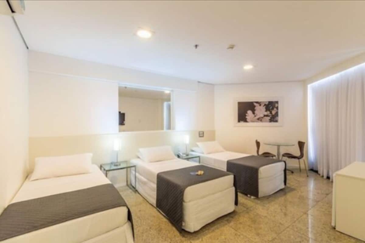 Habitación de hotel con dos camas, una mesita, una silla y un ventanal con cortinas.
