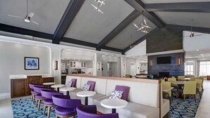 A spacious lobby with a high ceiling, modern lighting fixtures, a seating area with purple chairs and white tables, and a fireplace.
