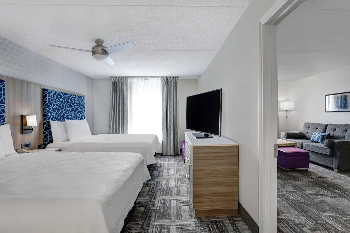 Ein Hotelzimmer mit zwei Betten, einem Flachbildfernseher, einem Schreibtisch, einer Couch und einem Deckenventilator.
