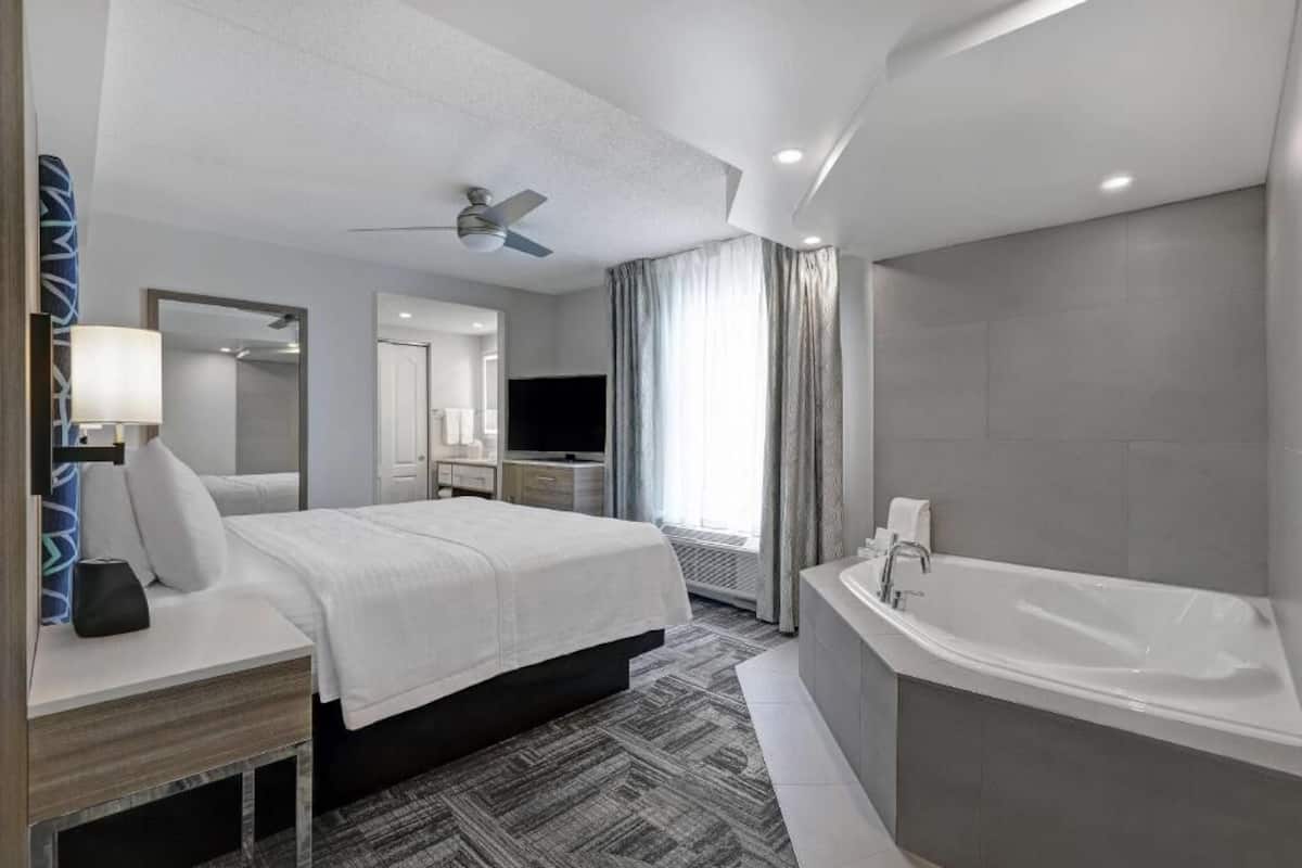 Ein modernes Hotelzimmer mit einem großen Bett, einem separaten Badezimmer inklusive Badewanne, einem Deckenventilator und einem Flachbildfernseher.