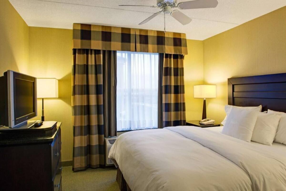 Ein Hotelzimmer mit einem Bett, zwei Nachttischlampen, einem Fernseher auf einem Schrank, einem Deckenventilator und einem Fenster mit Vorhängen.