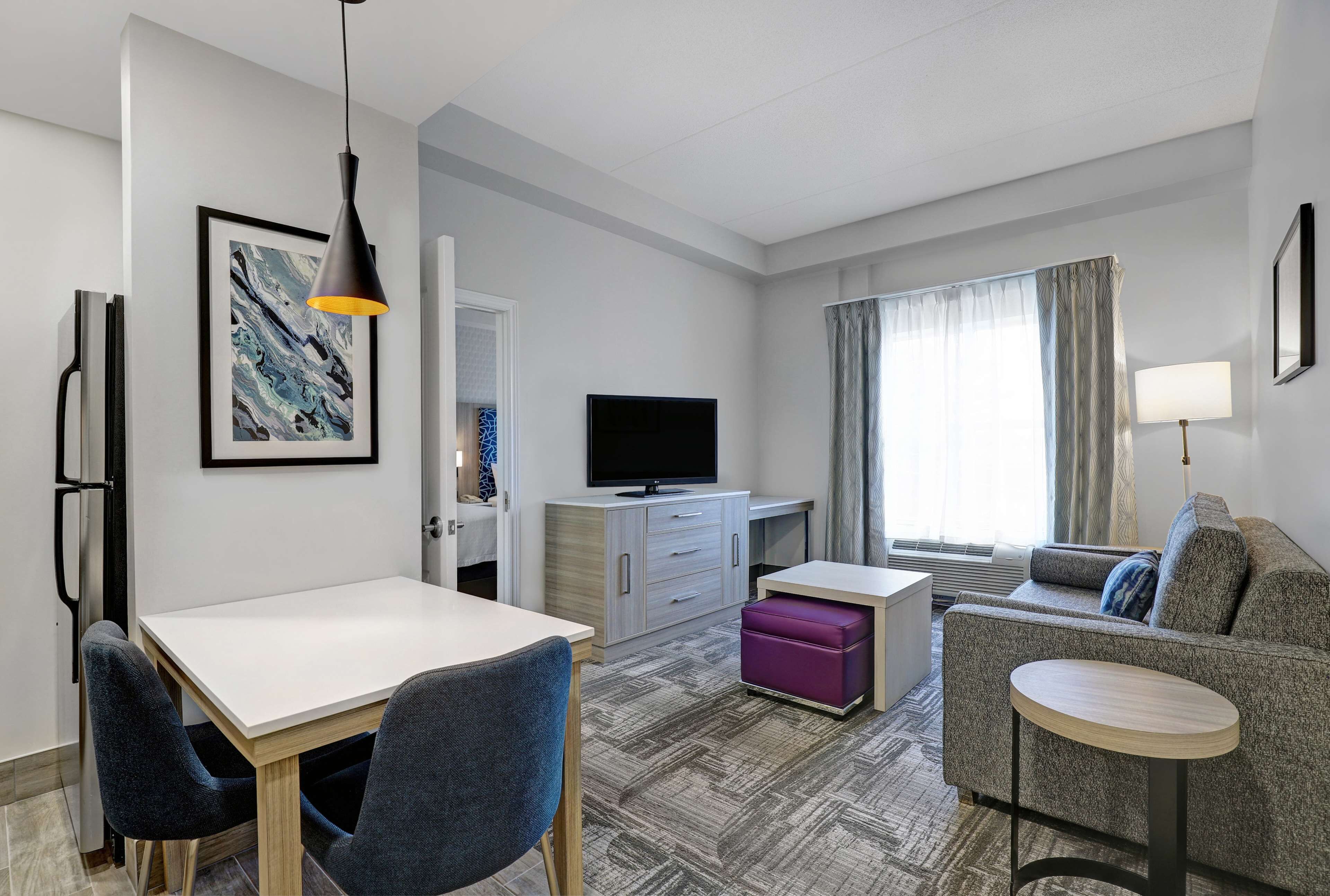 Una habitación de hotel moderna con zona de comedor, sofá, televisor y ventana con cortinas.