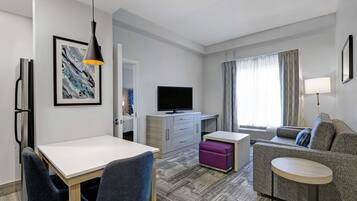 Una habitación de hotel moderna con zona de comedor, sofá, televisor y ventana con cortinas.