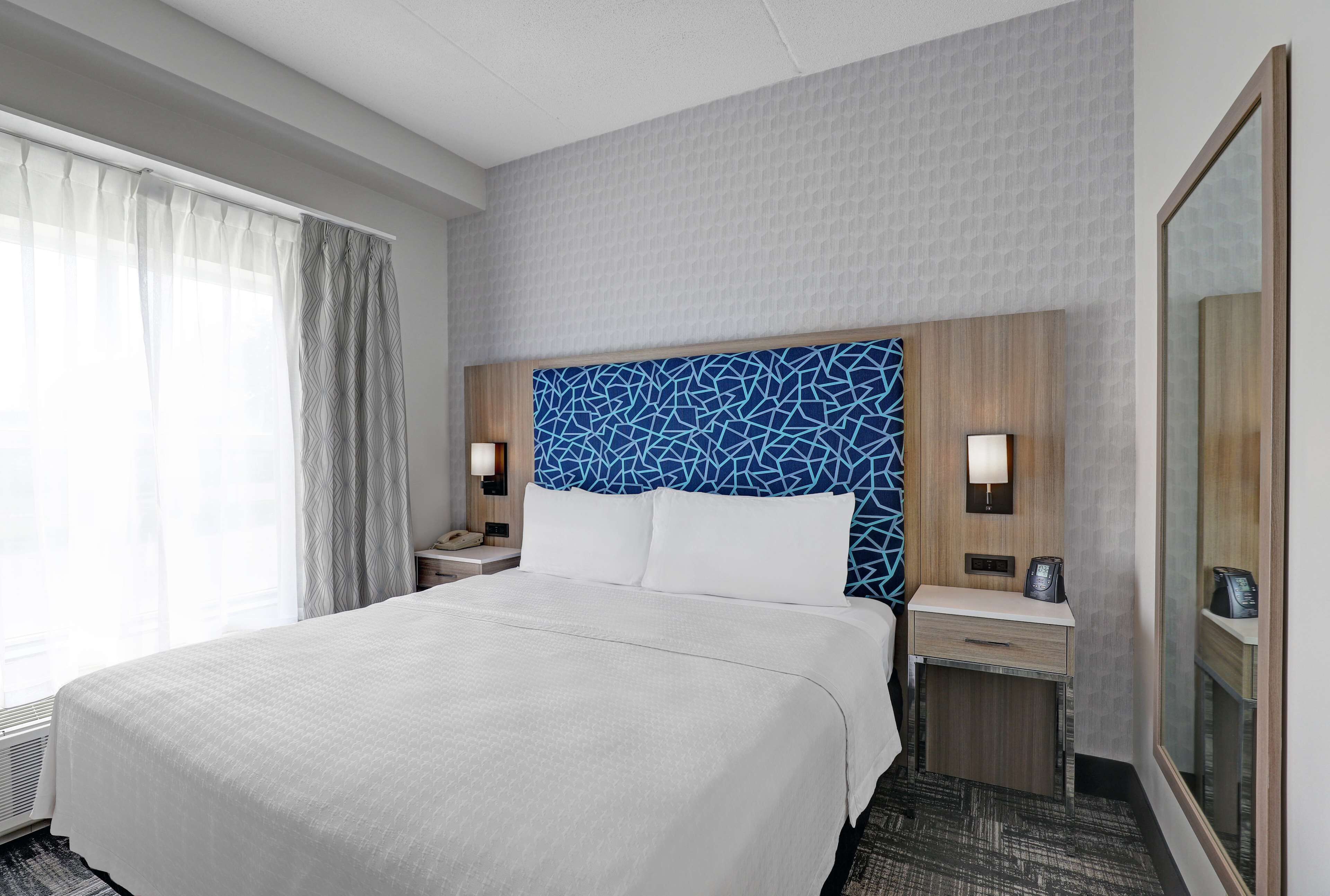Ett hotellrum med en stor säng, en sänggavel med ett blått, geometriskt mönster, två sängbord med lampor, en spegel och ett fönster med tunna, genomskinliga gardiner.