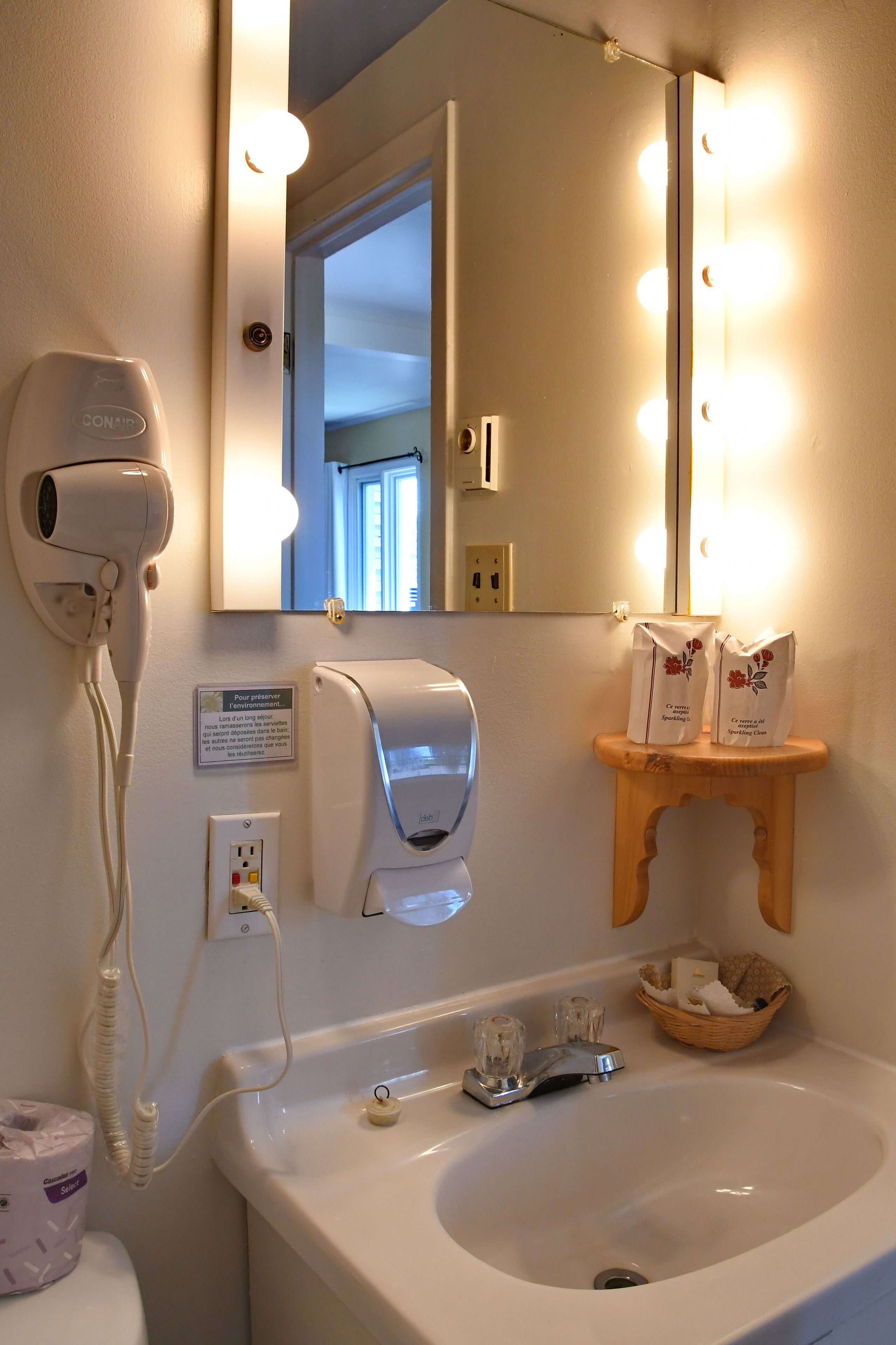 Une salle de bain équipée d’un sèche-cheveux, d’un sèche-mains, d’un miroir avec éclairage, d’un lavabo et d’un panier contenant des articles de toilette.