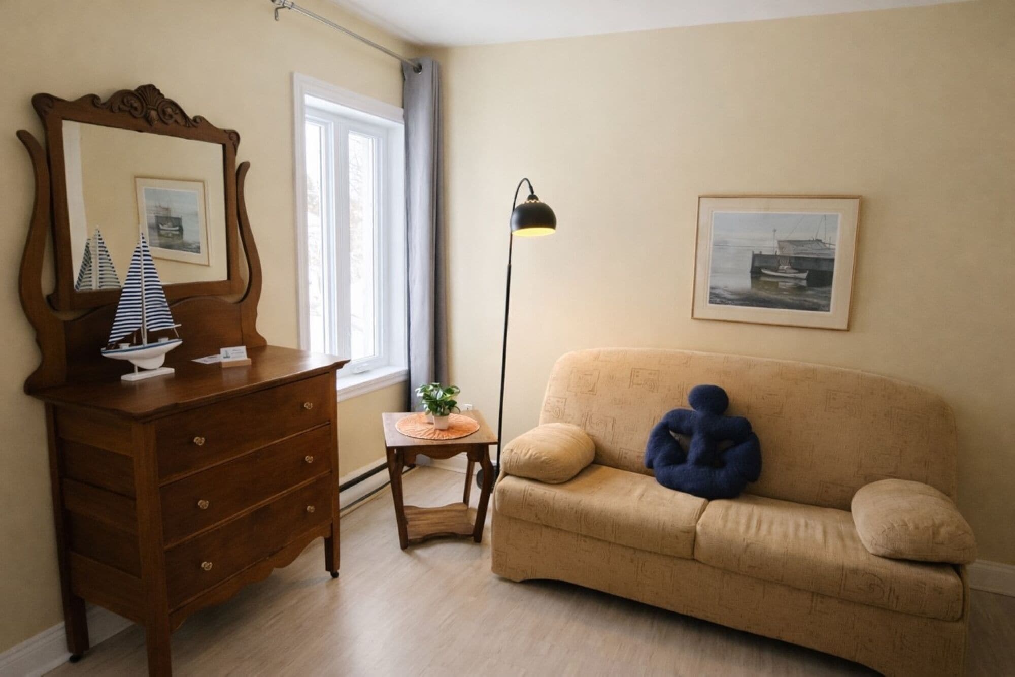 A room with a wooden dresser, a beige sofa, a floor lamp, and framed pictures on the walls.