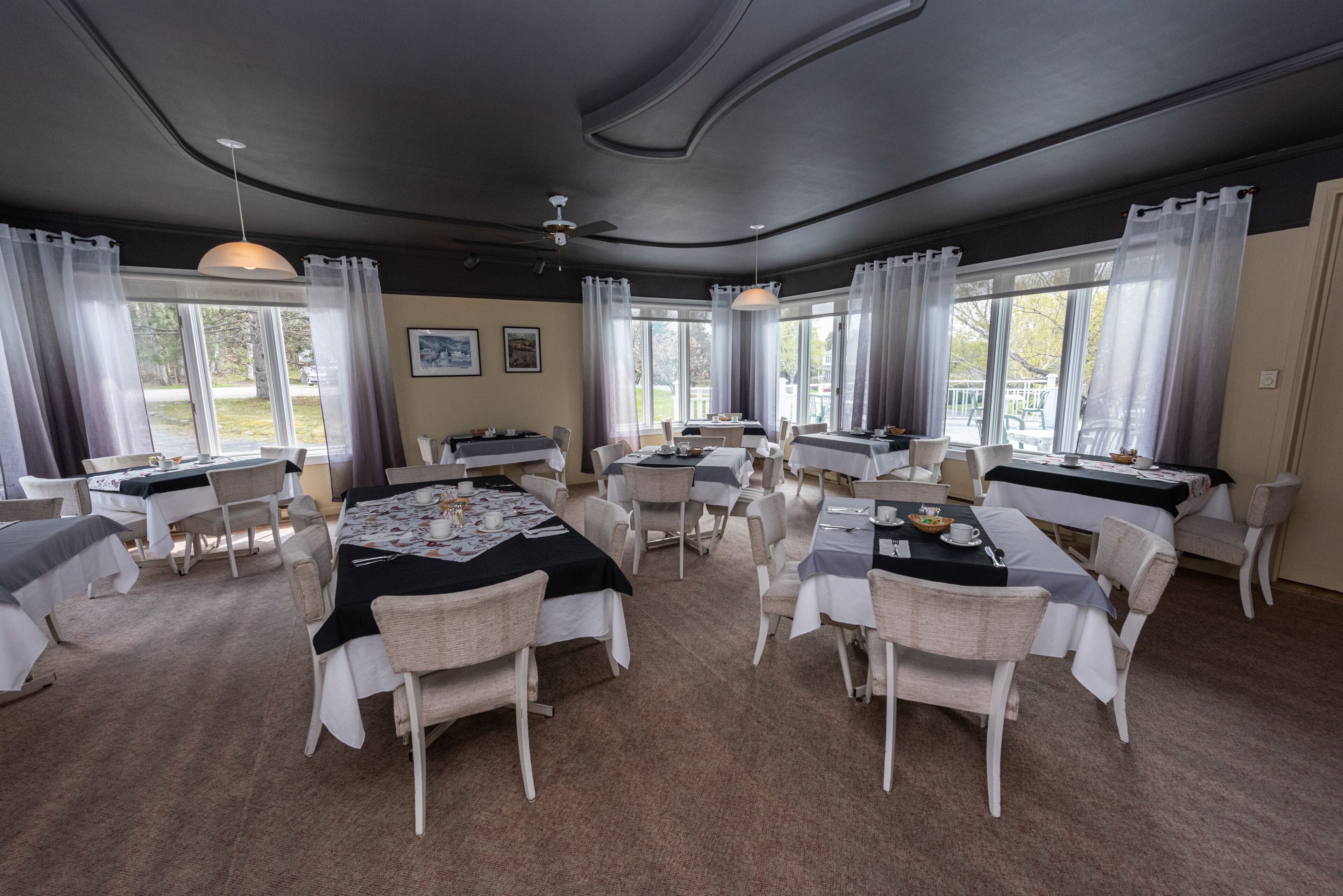 Un espace repas comprenant plusieurs tables et chaises, de grandes fenêtres avec des rideaux et un ventilateur de plafond.