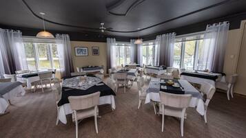 A dining area with multiple tables and chairs, large windows with curtains, and a ceiling fan.