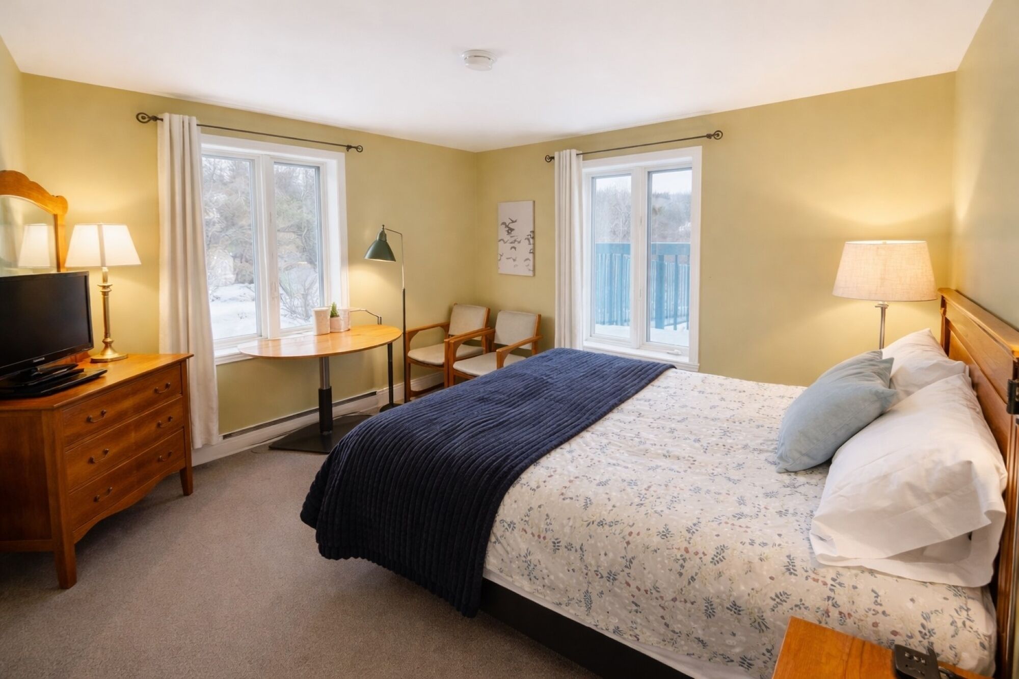 A bedroom with a bed, bedside table, lamp, TV, dresser, chair, and windows.