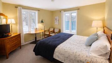 A bedroom with a bed, bedside table, lamp, TV, dresser, chair, and windows.