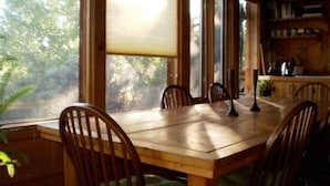 Une table à manger en bois, des chaises, de grandes fenêtres et un coin cuisine avec des étagères.