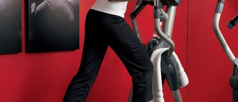 Una mujer usando una máquina elíptica en un gimnasio con paredes rojas y varias máquinas de ejercicio.