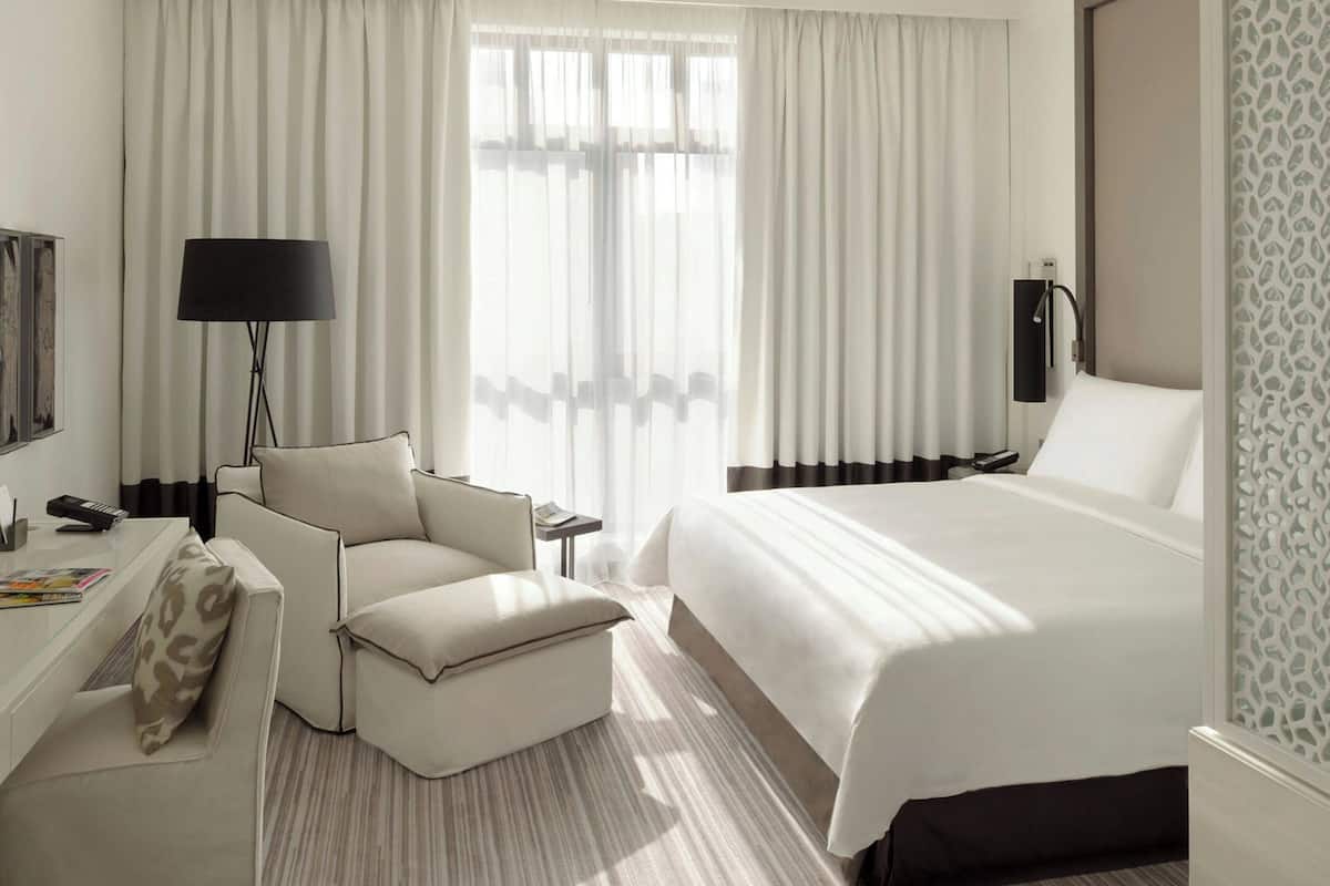 A hotel room with a bed, a chair, a desk, a lamp, and curtains.