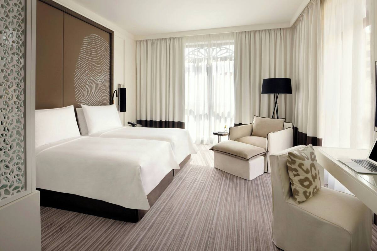 A hotel room with two beds, a desk with a laptop, a chair, a lamp, and a large window with curtains.
