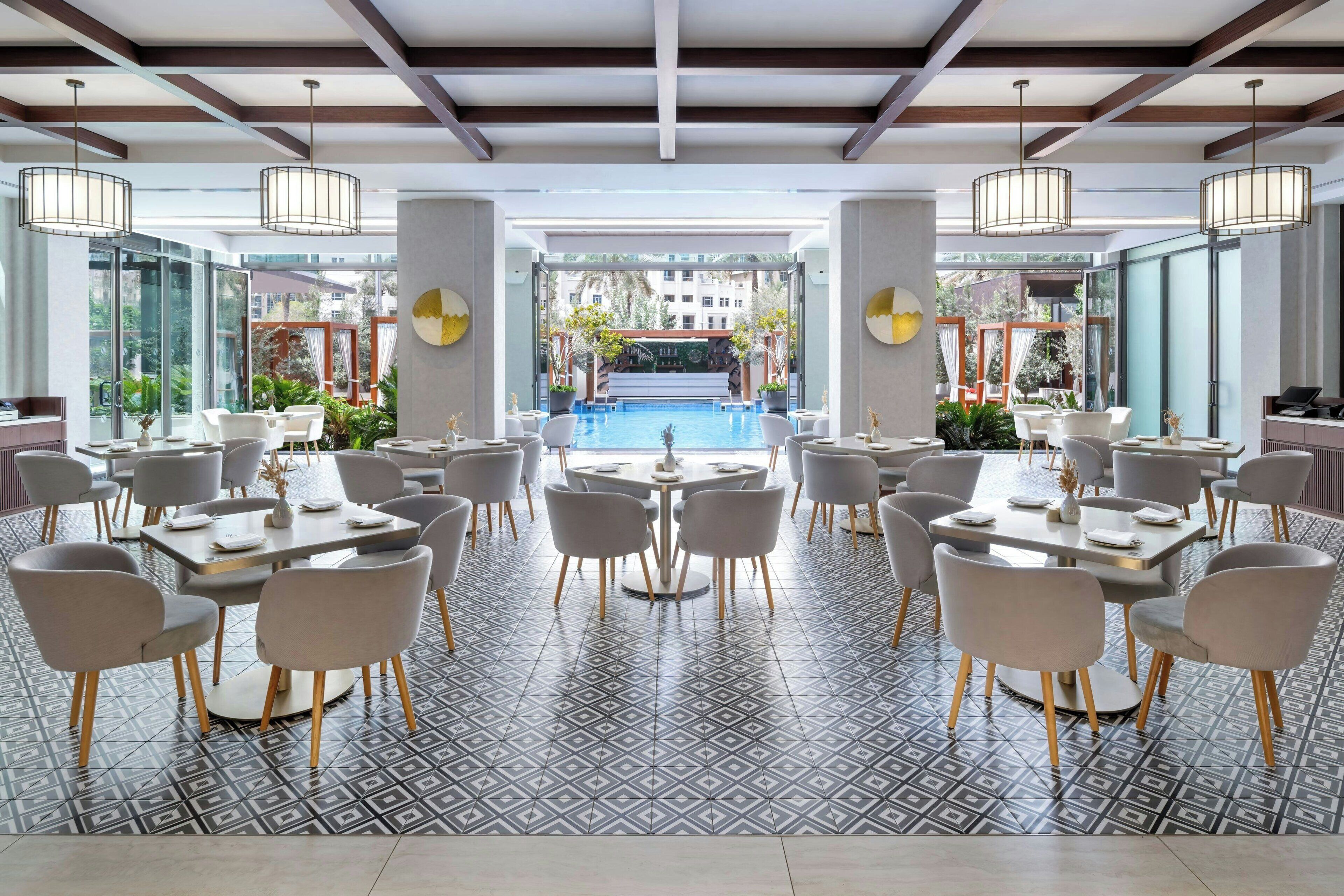 A spacious indoor dining area with multiple tables and chairs, a central pool, and large windows offering views of the surrounding area.