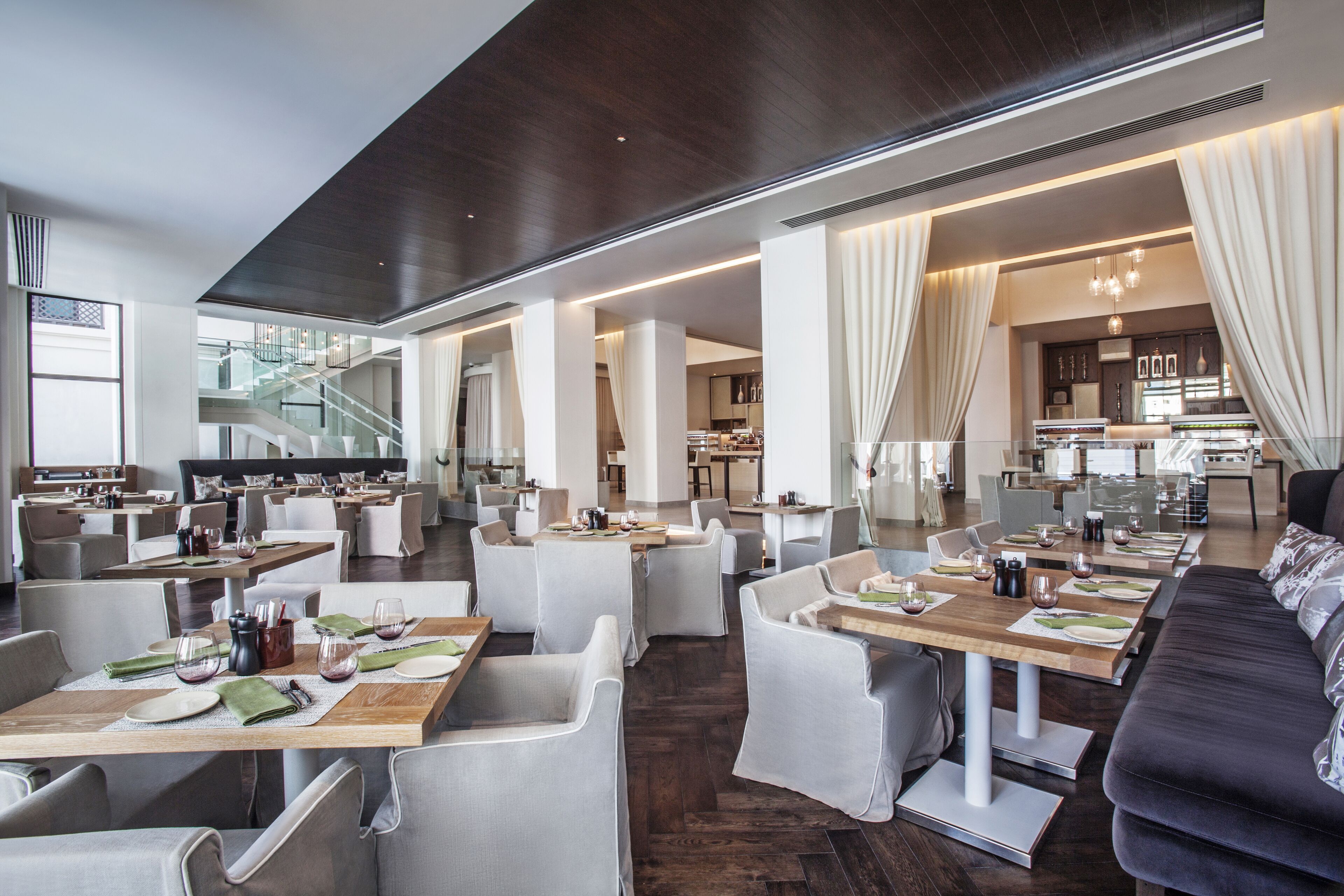 A spacious hotel dining area with multiple tables, chairs, and a modern kitchen in the background.